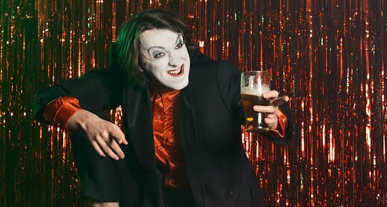 man with angry face, white makeup and green hair in front of red shimmering curtain