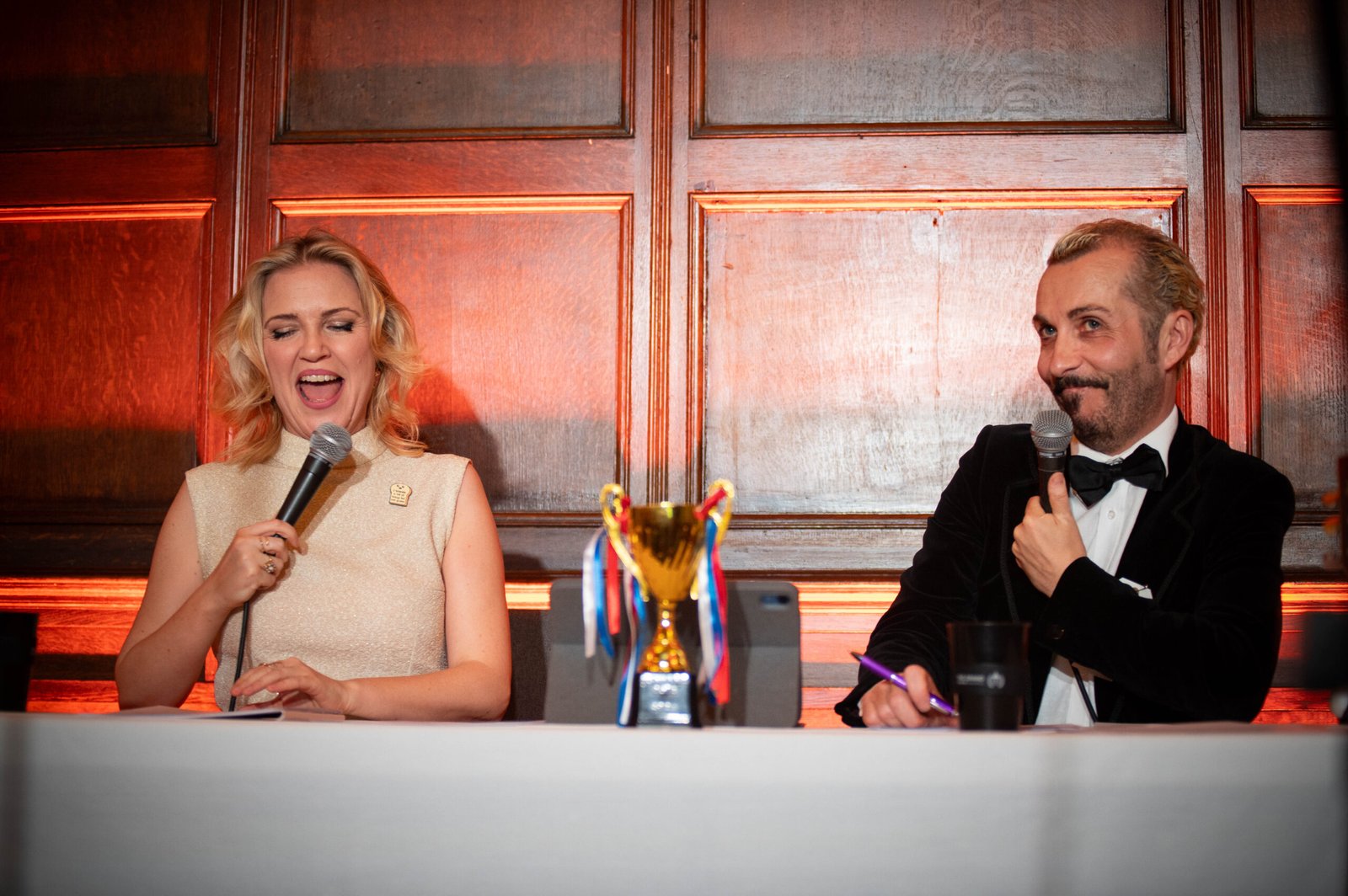 Female and male comedians laughing and between them is a gold trophy