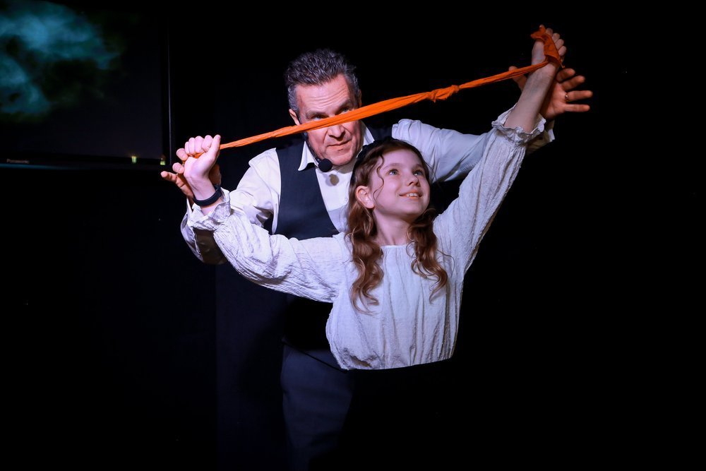 Male magician performs trick with scarf and a young girl on stage